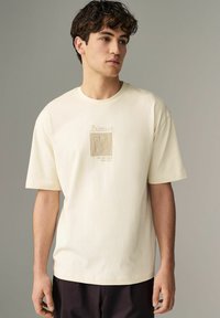 Cremefarbener Baumwoll-T-Shirt mit kurzen Ärmeln, versehen mit einem gestickten, von Picasso inspirierten Design auf der Brust in beiger Fadenfarbe.