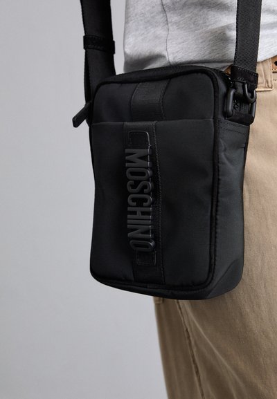 Svart nylon crossover-väska med präglad "MOSCHINO"-logotyp, justerbar rem, rektangulär form och dragkedjestängning.