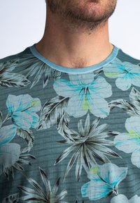 Teal t-paita lyhyillä hihoilla, jossa on kukkakuosi, sisältäen vaaleansinisiä ja harmaita hibiskuksia sekä vihreitä lehtiä korostamassa, ribbetty kaulus.