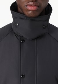 Black padded jacket featuring a high collar, snap buttons, and a smooth, matte texture. Close-up view of the collar and front details.