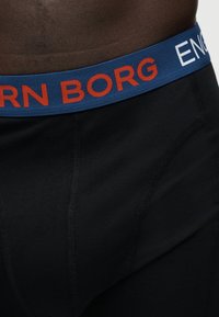 Μέση και άνω μηρός ατόμου που φοράει μαύρα μπόξερ με μπλε μέση με κείμενο «BJORN BORG» σε κόκκινο και λευκό.