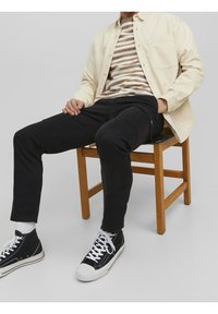 Ljusbeige skjorta med knappar över en randig brun och vit T-shirt, i kombination med slim-fit svarta byxor och svarta högst upp sneakers.