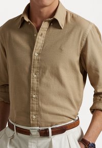 Camicia beige a maniche lunghe con bottoni e colletto, dotata di un piccolo logo. Tessuto casual, abbinata a una cintura in pelle marrone e pantaloni color crema.