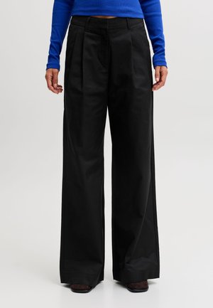 Personne portant un pantalon noir taille haute à jambes larges associé à un haut bleu à manches longues, debout devant un fond clair uni.