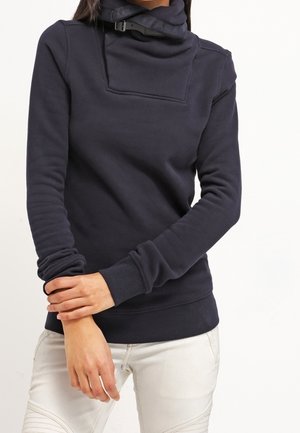 Pull-over bleu marine avec un col haut à rabat et un détail avec boucle en plastique. Comprend des manches longues ainsi que des poignets et un ourlet côtelés. Texture de tissu lisse.