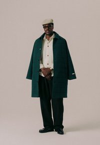 Grønn rutete frakk over en kremfarget skjorte med knapper, svarte bukser og mørke sko. En beige flat cap og briller fullfører antrekket.