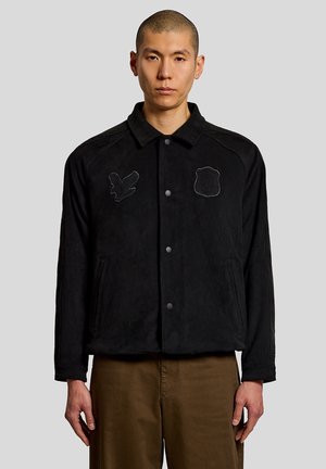 Mand iført en sort knappet jakke med to broderede patches og brune bukser, stående foran en ensartet grå baggrund.