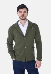 Blazer in maglia verde oliva con colletto a scialle, due tasche frontali, chiusura a bottone e un fazzoletto da taschino a fantasia. Il tessuto sembra testurizzato.