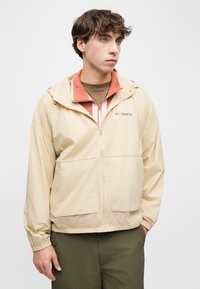 Νεαρός άνδρας φορώντας μπεζ αδιάβροχο Columbia πάνω από κόκκινο ζακέτα με φερμουάρ και χακί παντελόνι, στέκεται με ένα χέρι στ ην τσέπη, κοιτάζοντας αριστερά.