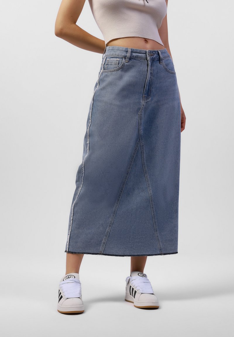 Vaaleansininen denim-midi-hame suoran leikkauksen, etusaumojen ja repaleisen helman kanssa. Yhdistetty valkoisiin lenkkareihin, joissa on mustat raidat.
