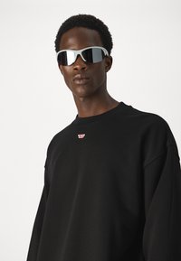 Črna trenerka s krožnim izrezom in diskretno logotipno podrobnostjo, združena z odsevnimi srebrnimi sončnimi očali, ki jih nosi oseba s kratkimi, kodrastimi lasmi.