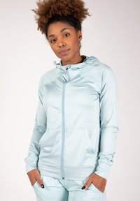 Haut de sport bleu clair avec zip intégral, capuche à cordon de serrage et poches kangourou ; texture lisse et design ajusté avec détail de logo.