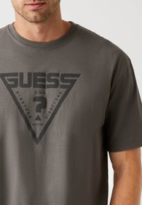 Hombre que lleva una camiseta gris con un gran logo negro de Guess que presenta un triángulo y un signo de interrogación en el pecho.