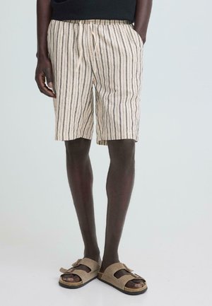Mann iført beige og svarte shorts med vertikale striper og beige sandaler med åpen tå, stående med hendene i lomma foran en ensfarget bakgrunn.