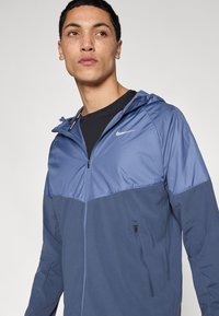 Μπλε μπουφάν με κουκούλα της Nike, διχρωμίας· διαθέτει φερμουάρ στο μπροστινό μέρος, πλαϊνές τσέπες και λογότυπο στο στήθος σε αντίθετη απόχρωση.