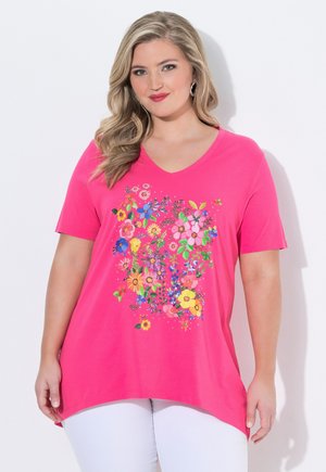 Kobieta o długich blond włosach, ubrana w jaskraworóżowy T-shirt z dekoltem w serek, z kolorowym kwiatowym nadrukiem i białe spodnie, stojąca na białym tle.