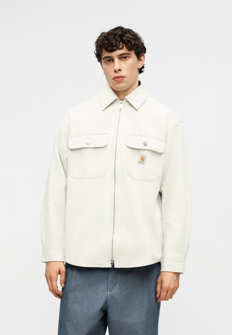 Junger Mann trägt eine hellbeige Corduroy-Jacke mit Reißverschluss und zwei aufgesetzten Taschen vorne, kombiniert mit lockeren blauen Jeanshosen.