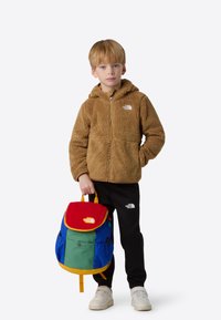 Rjava flis jopa z zadrgo, črne hlače in večbarven nahrbtnik z rdečim vrhom, modrim telesom in rumenimi poudarki. Bela obutev.