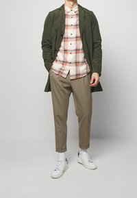 Groene lange jas over een geruite button-up-shirt in wit, oranje en marineblauw, gecombineerd met beige broek en witte sneakers.