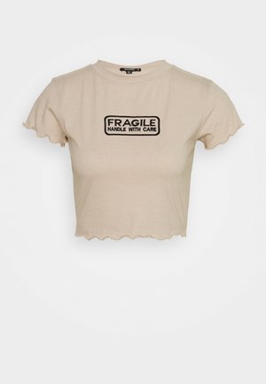 T-shirt beige court avec bords festonnés et texte en patch noir "FRAGILE HANDLE WITH CARE" centré sur le devant.