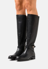 Stivali neri in pelle al ginocchio con cerniera laterale, cinturino regolabile con finiture dorate e suola piatta per il massimo comfort. Texture liscia.
