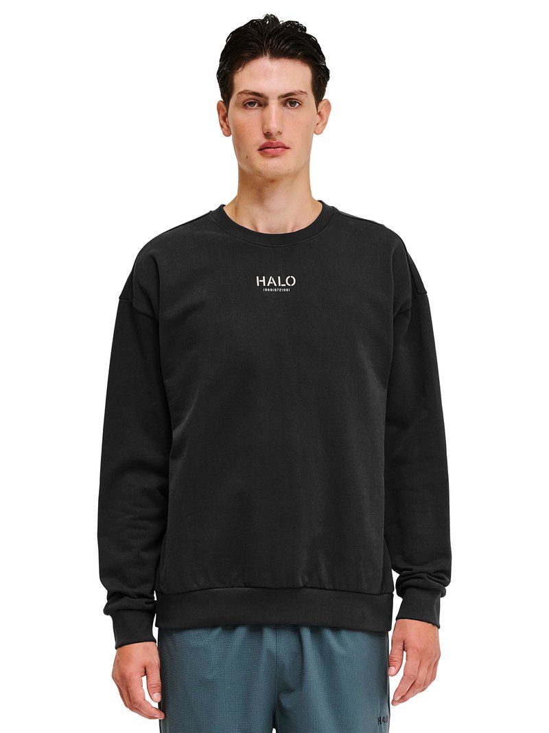 Man draagt een zwart sweatshirt met de tekst "HALO" en cijfers op de borst, gecombineerd met blauw gestructureerde broek, staand tegen een witte achtergrond.