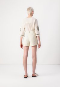 Personne aux cheveux blonds courts, vue de dos, portant un haut blanc brodé à manches longues, un short crème et des sandales plates beiges.