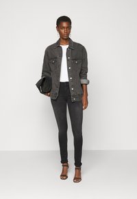 Jaqueta de denim cinza, blusa branca de canelado, calças skinny pretas e saltos altos pretos com tiras. Modelo segura uma mala preta. Design minimalista, estilo justo.