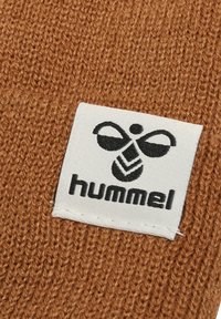 Brązowa dziana tkanina z białą metką z czarnym logo i napisem "hummel" poniżej. Tekstura jest miękka i wyrazista.
