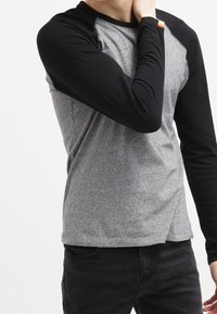 Person som bär en grå och svart långärmad tröja och med en hand som rör vid sin nacke, står framför en enfärgad vit bakgrund.