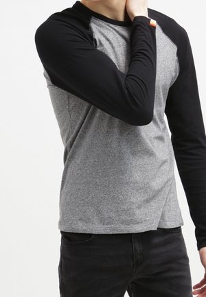 Person in einem grau-schwarzen Langarmshirt, die mit einer Hand den Hals berührt, vor einem schlichten weißen Hintergrund stehend.