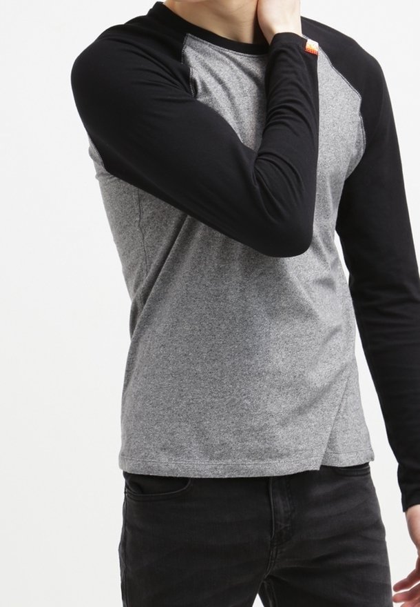 Person som bär en grå och svart långärmad tröja och med en hand som rör vid sin nacke, står framför en enfärgad vit bakgrund.