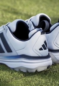 Paire de chaussures de golf blanches et noires avec semelles crantées placées sur de l'herbe verte, montrant les vues arrière et latérales.