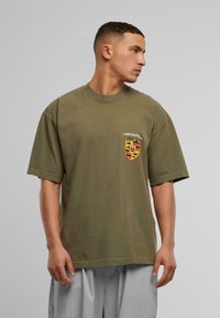 Olivenfarvet bomulds t-shirt med rund hals, korte ærmer og et skjolddesign på venstre bryst med gule, sorte og røde detaljer.