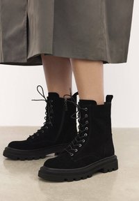 Botines de ante negros con cordones en la parte delantera y cremallera lateral. Suela de goma gruesa con superficie texturizada y lengüeta en la parte trasera.