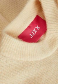 Pull en tricot de couleur crème montrant une étiquette de vêtement rouge avec le texte blanc "JUXX" à l'intérieur du col.