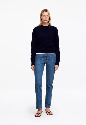 Femme aux cheveux blonds raides portant un pull noir à manches longues, un jean bleu et des sandales noires à bout ouvert, debout devant un fond blanc.