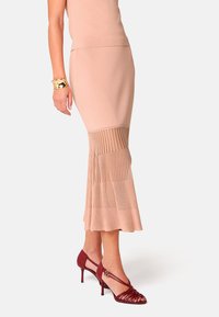 Ribbel gebreide rok tot op de knie in lichtroze, met een uitlopende zoom; gecombineerd met bordeauxrode hakken met gekruiste banden en gespen.