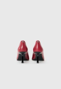 Red leather high-heeled shoes with a pointed toe and tapered silhouette. Features a black heel and metallic logo detail at the back.