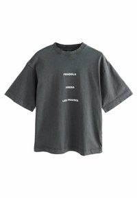 Camiseta negra de gran tamaño hecha de algodón con mangas cortas. En el frontal presenta texto blanco: “FRAGOLA,” “FRESA,” “LES FRAISES,” alineado verticalmente.