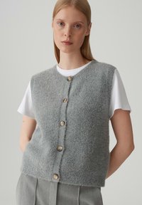Gilet in misto lana grigio chiaro con scollatura tonda, caratterizzato da cinque bottoni in legno e una superficie morbida e testurizzata. Indossato sopra una camicia bianca.