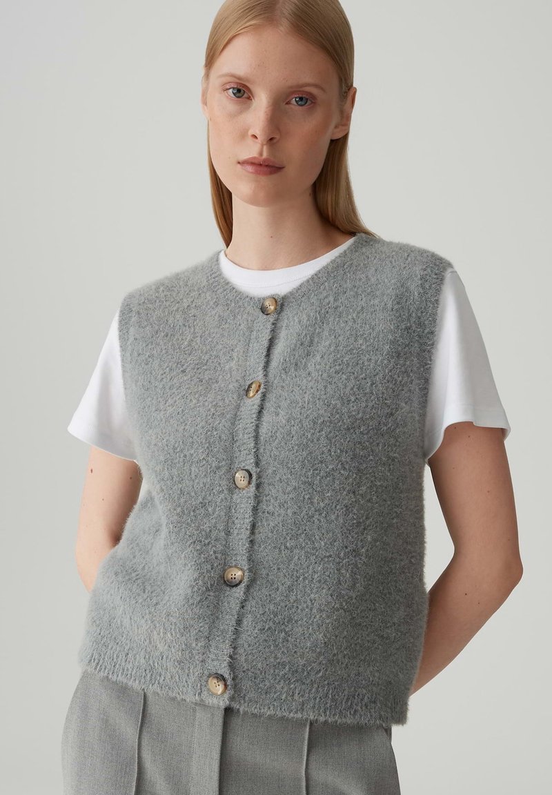 Gilet in misto lana grigio chiaro con scollatura tonda, caratterizzato da cinque bottoni in legno e una superficie morbida e testurizzata. Indossato sopra una camicia bianca.