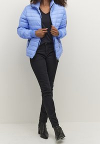 Mujer vistiendo una chaqueta acolchada azul claro, camiseta negra de cuello en V, jeans ajustados negros y botas de tobillo negras con tacón, de pie sobre un suelo blanco.