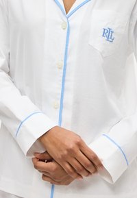 Λευκό πουκάμισο με κουμπιά, με γαλάζια διακόσμηση, κεντημένο "RLL" στην τσέπη στο στήθος, φορεμένο από άτομο με τα χέρια ενωμένα κοντά στη μέση.