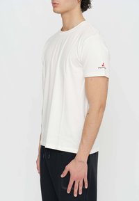 T-shirt blanc à manches courtes en coton, avec un col rond et un petit logo rouge sur la manche gauche. Texture lisse, coupe régulière.