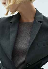 Blazer sur mesure gris foncé avec une texture subtile, un bouton unique et une poche poitrine, superposé à un pull gris clair.
