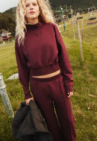 Bordová krátka mikina s vysokým golierom a dlhými rukávmi v kombinácii so zodpovedajúcimi širokými nohavicami. Modelka drží čiernu bundu. Pozadie z čerstvej zelenej trávy.