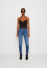 Top halter nero con bordi arricciati, abbinato a jeans blu aderenti e stivali neri alla caviglia. Modella che indossa occhiali da sole neri.