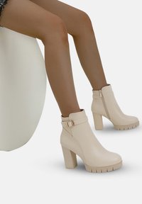 Botas de tobillo beige con acabado brillante, tacón de bloque, cremallera lateral y detalle de hebilla, con una suela texturizada para proporcionar agarre.