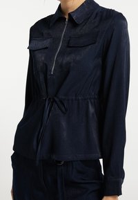 Marineblauwe shirt met een kraag, ritssluiting en twee voorzakken. Heeft een gathered taille met een trekkoord en een gladde textuur.